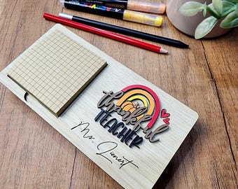May include: A wooden desk organizer with a notepad and a decorative rainbow design that says "thankful TEACHER" and "Ms. Juenert". The organizer also has a red pencil, a black pencil, and two markers.