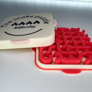 May include: A white and red snake puzzle. The lid is printed with "the snake puzzle" and "Difficulty". The open puzzle reveals a red maze within a white frame. The lid features a smiling face.