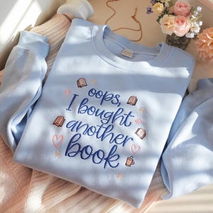 Può includere: Una felpa azzurra con la scritta ricamata "oops, I bought another book" in blu navy. Il design include motivi ricamati di libri e cuori in rosa e marrone chiaro, e una piccola tazza di caffè.