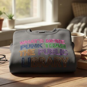 May include: A dark gray sweatshirt with the embroidered text "WHAT'S MORE PUNK THAN THE PUBLIC LIBRARY" in various colors. The sweatshirt is folded on a wooden surface, with a coffee cup and books in the background.