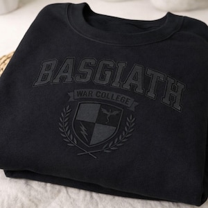 Pode incluir: Camisola preta com o texto "BASGIATH" na parte superior e "WAR COLLEGE" abaixo. Um brasão com um dragão, um raio e uma coroa de louros está centrado abaixo. A camisola é feita de um material macio.