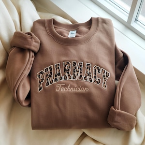 Peut inclure: Un sweat-shirt marron avec le mot "PHARMACY" en grandes lettres à imprimé léopard sur la poitrine. Le mot "Technician" est brodé en dessous en plus petit, en écriture cursive. Les manches sont retroussées.
