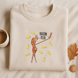 Puede incluir: Sudadera de color crema con una ilustración de dibujos animados de un personaje de pasta rigatoni con los brazos extendidos, rodeado de formas de pasta rigatoni amarillas. El texto "RIGATONI PASTA" está encima del personaje.