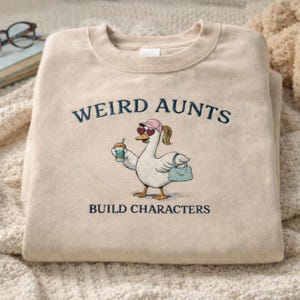 May include: A beige sweatshirt with the embroidered text "WEIRD AUNTS BUILD CHARACTERS". The graphic shows a cartoon goose wearing sunglasses, a pink cap, and carrying a purse and a drink. The sweatshirt is folded.