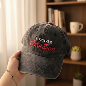 Puede incluir: Gorra de béisbol gris con el texto bordado "i need a Coke Zero" en rojo. La gorra está sujeta por una persona y tiene una visera curva. El sombrero es un accesorio informal.