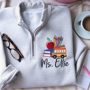 Puede incluir: Sudadera gris claro con cuello con cremallera. Presenta un diseño bordado de una manzana, libros, crayones y un autobús escolar. El texto "Ms. Ellie" está debajo. También se ven gafas y una taza de café.