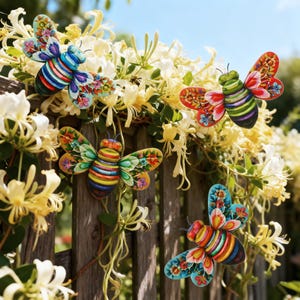 May include: Four decorative metal bee ornaments with floral wing patterns are attached to a wooden fence. The bees feature striped bodies in various colours, including blue, green, red, and orange. White and yellow flowers are in the background.