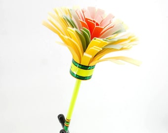 May include: A colourful paper flower with a yellow stem and a multi-coloured flower head. The flower head features layers of yellow, green, orange, pink, and white paper. The stem is light green with green and yellow stripes near the base.