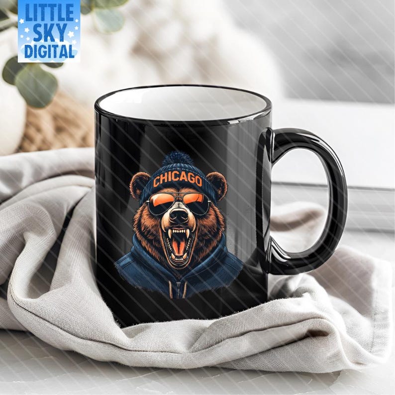 Black ceramic coffee mug featuring a roaring bear graphic wearing a navy blue beanie with orange “CHICAGO” text, orange aviator sunglasses, and a blue hoodie. The mug sits on light gray fabric atop a marble surface with white and gray veining. Background includes blurred green eucalyptus leaves and soft white decor, creating a cozy, stylish setting ideal for product mockups or lifestyle listings.