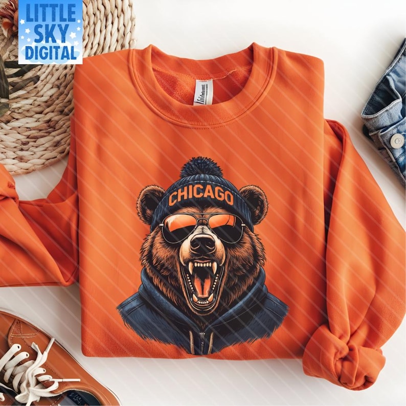 Folded orange sweatshirt featuring a bold graphic of a roaring bear wearing a dark blue beanie with “CHICAGO” in orange text, orange aviator sunglasses, and a blue hoodie. Displayed on a white surface with orange sneakers, denim clothing, and a straw placemat with sunglasses, creating a vibrant, sporty flat lay ideal for showcasing apparel.
