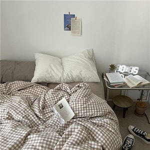 May include: A bed with a brown and white checkered duvet cover and two white striped pillows. A small side table holds a digital clock, books, and a small plant. A pair of black and white sneakers sits on the floor.