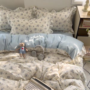 May include: A bed with a light blue duvet and floral patterned bedding. The pillows have ruffled edges. A small teddy bear with a red heart and a pair of gray headphones rest on the bed. A vintage keyboard is at the foot of the bed.