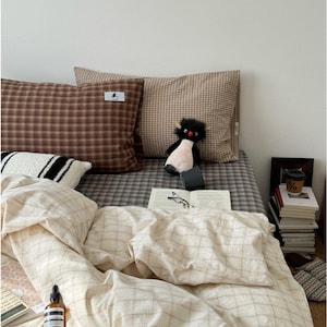 Op de afbeelding: Een bed met een crème en bruin geruit dekbedovertrek en kussenslopen. Een knuffel pinguïn zit op het bed naast een open boek en een bril. Een fles serum en een stapel boeken zijn ook zichtbaar.