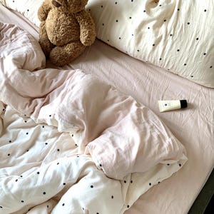 May include: A bed with a light pink sheet and a white duvet cover with black polka dots. A brown teddy bear sits on the bed, and a small tube of lotion rests on the sheet. The bedding is rumpled, suggesting use.