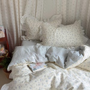 Puede incluir: Una cama con ropa de cama con estampado floral, que incluye almohadas y un edredón. Las almohadas tienen bordes con volantes. La funda nórdica es de color crema claro con pequeñas flores azules. Un libro descansa sobre la cama. Cortinas con estampado floral en el fondo.