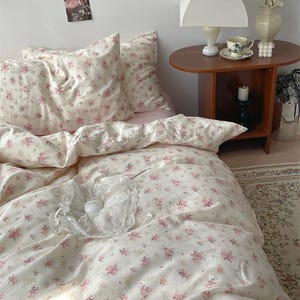 Pode incluir: Uma cama com uma colcha e fronhas com estampas florais em creme e rosa. A roupa de cama é adornada com um padrão delicado e repetitivo de pequenas flores rosa. Um acessório de renda branca repousa sobre a colcha. A cena é ambientada em um quarto com uma mesa lateral.