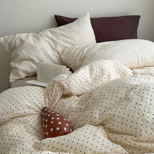 Może przedstawiać: Łóżko z kremową pościelą w małe czarne kropki. Widoczne są dwie poduszki z tym samym wzorem, a także ciemnobordowa poduszka. Brązowy przedmiot dekoracyjny z białymi kropkami spoczywa na łóżku, w pobliżu otwartej książki.