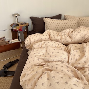 May include: A bed with a brown fitted sheet, pillows, and a duvet cover. The duvet cover and pillowcases are a light beige color with a repeating pattern of small gray floral designs. A small side table with a lamp and books is visible.