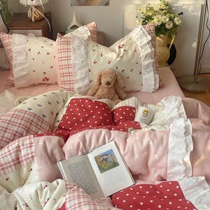 Könnte beinhalten: Ein Bett mit einer rosa Bettdecke und Kissen. Die Kissen haben Kirsch- und Karomuster mit weißen Rüschen. Ein Teddybär und ein offenes Buch liegen auf dem Bett. Die Bettwäsche hat ein rotes Tupfenmuster.