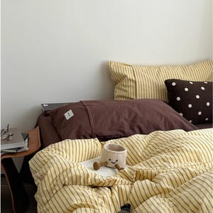 以下が含まれることがあります： 黄色とグレーのストライプの掛け布団と枕カバーが特徴のベッド。茶色の枕と白い水玉模様の茶色のクッションもベッドにあります。笑顔の顔が付いた小さな織りバスケットが掛け布団の上にあり、本とボトルも置かれています。