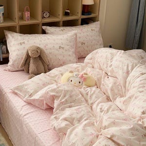 Puede incluir: Una cama con ropa de cama rosa con un pequeño estampado floral y de conejitos. El juego incluye una sábana bajera, fundas de almohada y un edredón. Un conejito de peluche y un peluche de Hello Kitty están en la cama, creando un ambiente acogedor.