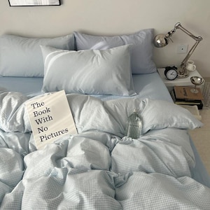 May include: A bed with light blue gingham bedding, including pillows and a duvet. A book titled "The Book With No Pictures" rests on the bed. A clear glass bottle and a silver desk lamp are also visible.