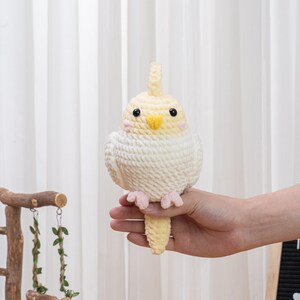 May include: A handcrafted crochet bird in cream and yellow, with black eyes and pink feet. The bird is held in a hand, highlighting its detailed craftsmanship. The bird's crest and beak are yellow, and it has a textured, fluffy appearance.