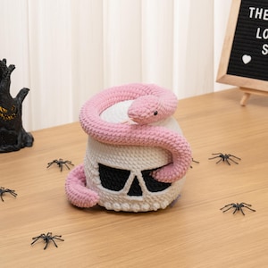 Puede incluir: Una calavera de ganchillo blanca con cuencas oculares y cavidad nasal negras, con una serpiente de ganchillo larga y texturizada de color rosa claro enroscada alrededor de la parte superior y los lados. La serpiente tiene ojos negros pequeños.