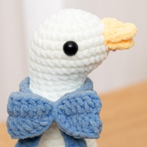 May include: A white crocheted plush goose toy featuring a soft, textured yarn construction. The goose has a bright yellow beak, a single round black plastic eye, and is wearing a prominent, textured blue crocheted bow tie around its neck.