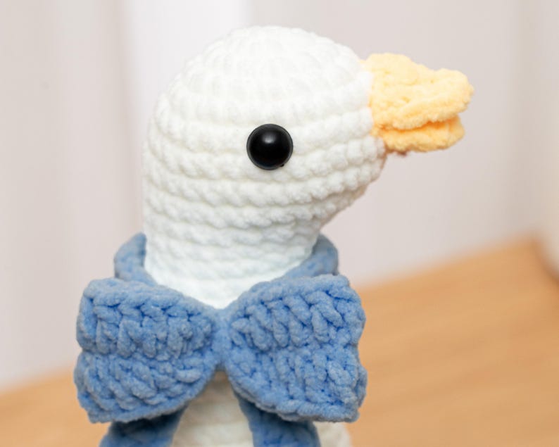 May include: A white crocheted plush goose toy featuring a soft, textured yarn construction. The goose has a bright yellow beak, a single round black plastic eye, and is wearing a prominent, textured blue crocheted bow tie around its neck.