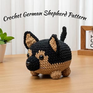 Può includere: Un cane da pastore tedesco all'uncinetto in nero e marrone chiaro. Il peluche fatto a mano ha grandi orecchie dritte e una piccola coda. Il testo "Crochet German Shepherd Pattern" è sopra il cane. Il cane è su una superficie di legno.