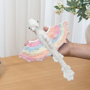 May include: A crocheted dragon toy featuring a white body and head with large, textured wings displaying a pastel rainbow gradient of pink, yellow, and blue yarn. The creature has a long, segmented tail and small black eyes, with a small tag reading The Loop Story.