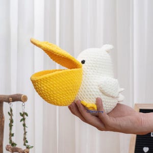 May include: A handmade, white crochet pelican with a large, open yellow beak. The bird has small black eyes and is held in a person's hand. The pelican's body is textured, and the beak is a bright yellow color. The overall design is soft and whimsical.