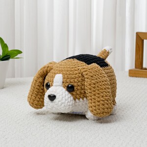 May include: A crocheted beagle dog plushie in shades of brown, black, and white. The handmade stuffed animal has large floppy ears, a white muzzle, and black eyes. The dog is lying down on a white surface.