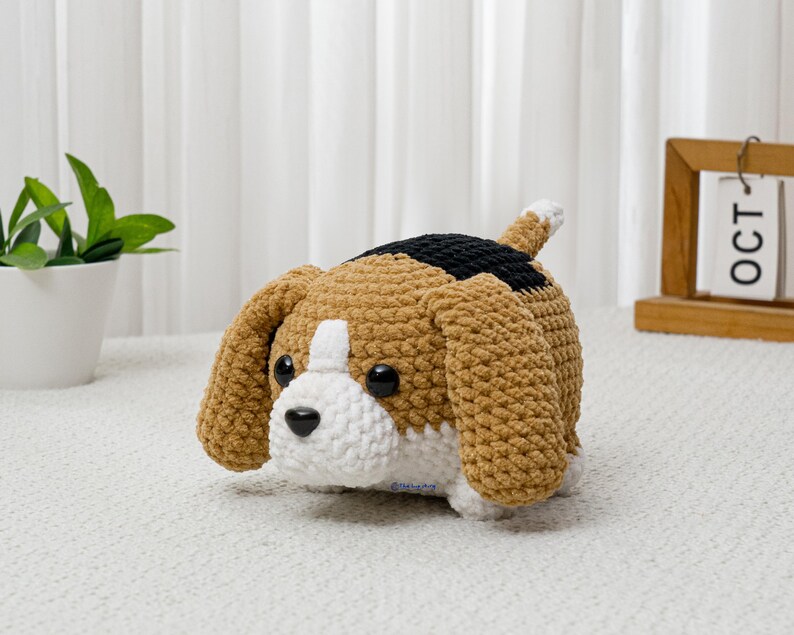 May include: A crocheted beagle dog plushie in shades of brown, black, and white. The handmade stuffed animal has large floppy ears, a white muzzle, and black eyes. The dog is lying down on a white surface.