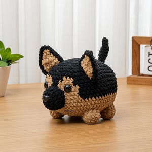 May include: A crocheted German Shepherd dog figurine in black and tan. The handmade plush toy has a rounded body, small legs, and upright ears. The dog is sitting on a wooden surface, with a small plant and calendar in the background.