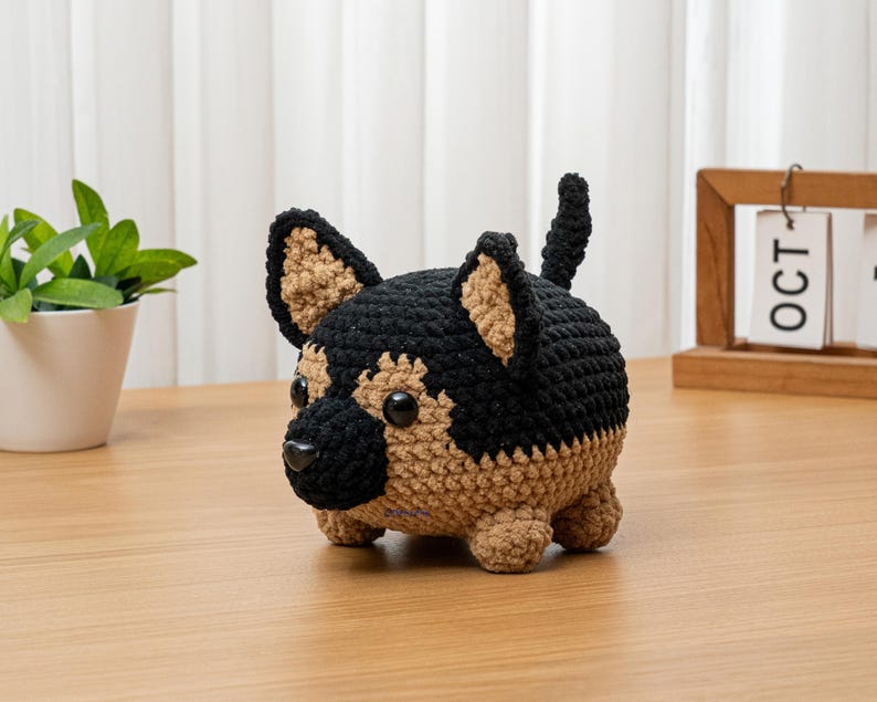 May include: A crocheted German Shepherd dog figurine in black and tan. The handmade plush toy has a rounded body, small legs, and upright ears. The dog is sitting on a wooden surface, with a small plant and calendar in the background.