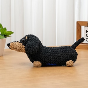 May include: A crocheted dachshund dog figurine in black, tan, and beige. The handmade dog has a long body, short legs, and a small tail. The dog is sitting on a wooden surface.