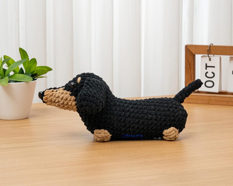 May include: A crocheted dachshund dog figurine in black, tan, and beige. The handmade dog has a long body, short legs, and a small tail. The dog is sitting on a wooden surface.