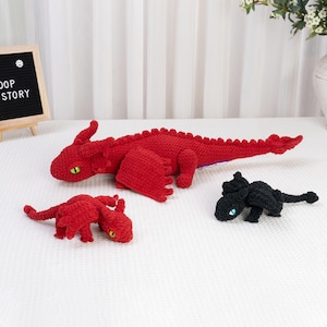 Könnte beinhalten: Drei gehäkelte Drachen-Stofftiere. Ein großer roter Drache mit grünen Augen, ein kleiner roter Drache mit gelben Augen und ein kleiner schwarzer Drache mit blauen Augen. Jeder Drache hat strukturiertes Garn, Flügel und einen langen Schwanz.