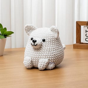 May include: A handmade, white crocheted bear figurine with black eyes and a black nose. The bear has small, rounded ears and is sitting on a wooden surface. A small green plant in a white pot is to the left of the bear.