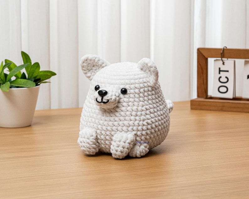 May include: A handmade, white crocheted bear figurine with black eyes and a black nose. The bear has small, rounded ears and is sitting on a wooden surface. A small green plant in a white pot is to the left of the bear.