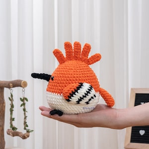 May include: A handmade crochet bird in orange, white, and black, featuring a spiky crest, black beak, and patterned wing. The bird is held in a hand. This is a whimsical, handcrafted item.