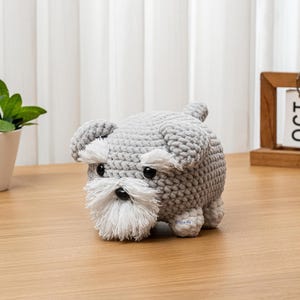 May include: A handmade gray crochet dog with white accents on the face and a small tail. The dog has black eyes and a white beard. The dog is sitting on a wooden surface.