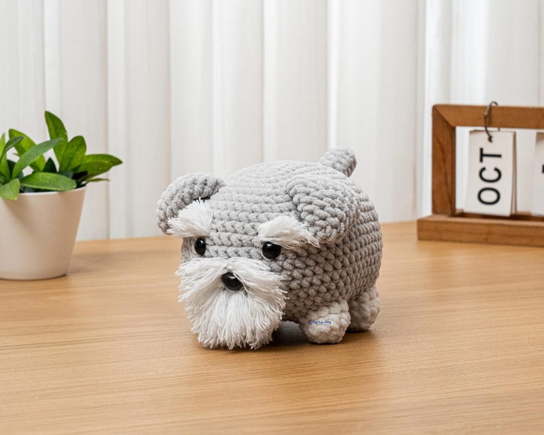 May include: A handmade gray crochet dog with white accents on the face and a small tail. The dog has black eyes and a white beard. The dog is sitting on a wooden surface.