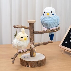 Op de afbeelding: Twee gehaakte vogelbeeldjes, één wit en geel, de andere blauw en wit, op een houten standaard. De standaard is gemaakt van takken en een houten basis. De vogels hebben zwarte ogen en zitten op takken.