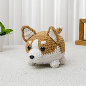 May include: A crocheted corgi dog plushie in brown and white. The handmade stuffed animal has a rounded body, small legs, and large ears. The corgi has black eyes and a black nose. The plushie is sitting on a white surface.