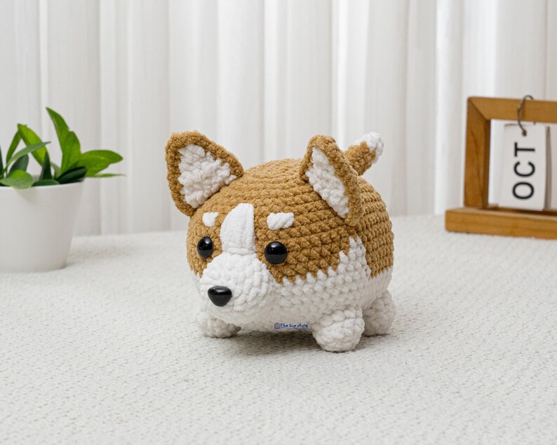 May include: A crocheted corgi dog plushie in brown and white. The handmade stuffed animal has a rounded body, small legs, and large ears. The corgi has black eyes and a black nose. The plushie is sitting on a white surface.
