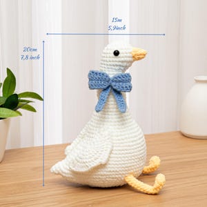 May include: A white crocheted goose plush toy featuring a light blue bow around its neck and yellow feet. The goose has black bead eyes and an orange beak. Dimensions indicated on the image are 7.8 inches in height and 5.9 inches in width.