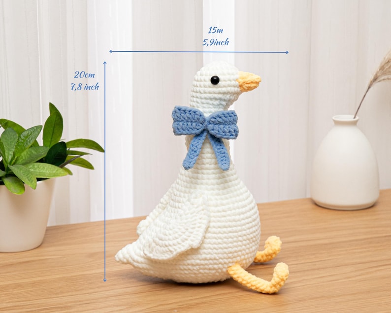 May include: A white crocheted goose plush toy featuring a light blue bow around its neck and yellow feet. The goose has black bead eyes and an orange beak. Dimensions indicated on the image are 7.8 inches in height and 5.9 inches in width.